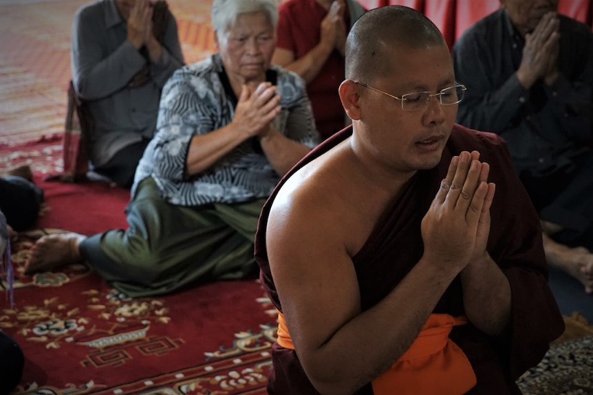 A monk in the foreground leading prayers and an elderly lady sitting behind him on the ground