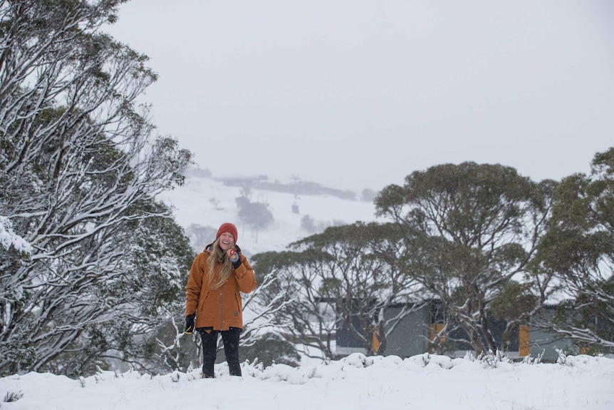 A smiling woman stands in thick snow on a mountain-side, holding a snow ball.