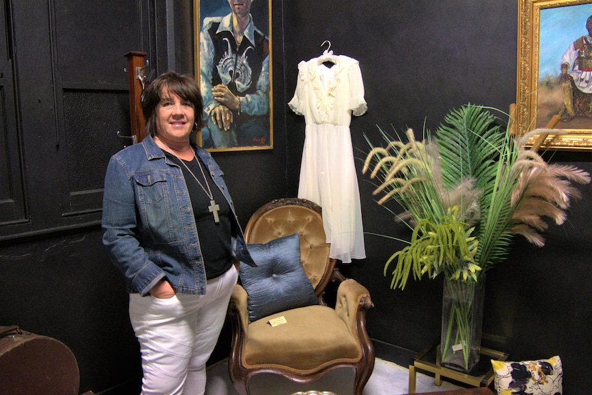 A woman stands smiling next to a vintage chair, framed art and a white dress hanging on the wall.