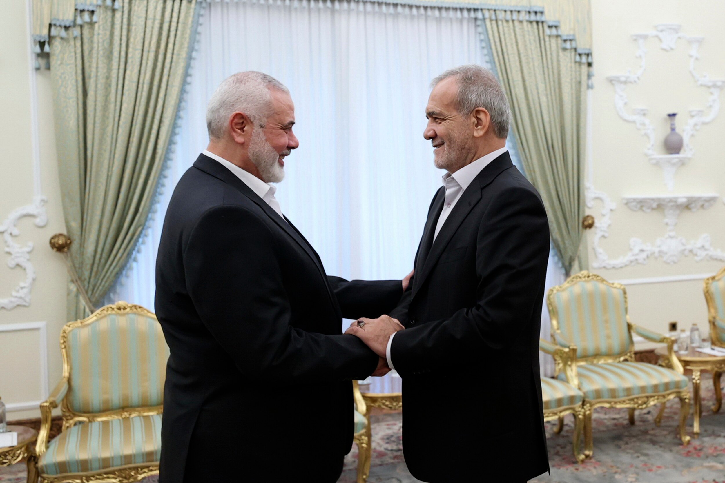 Masoud Pezeshkian shakes hands with Ismail Haniyeh as both men smile and stand in a sparse formal meeting room.