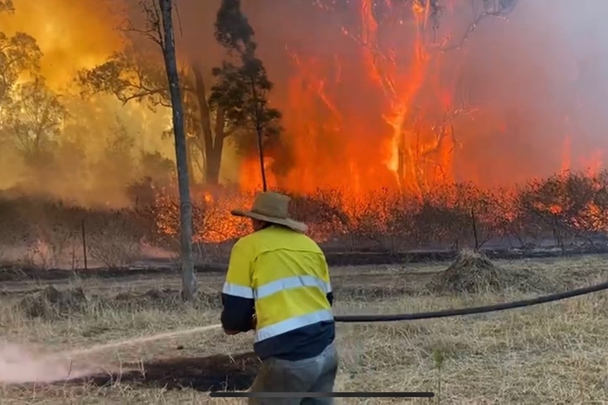 A man wearing hi-vis and a hat with a fire hose at a bush fire. 