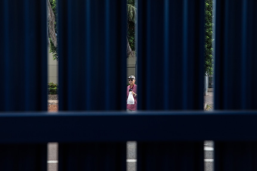 through the gaps of a fence, a man can be seen walking down a street.