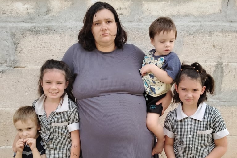 A woman in a purple dress with dark brown hair looks defeatedly at the camera, she has four children around her.