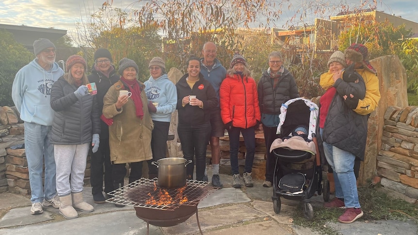 group of older people in warm clothes haddling around a fire in a stone paved area with trees and building in background