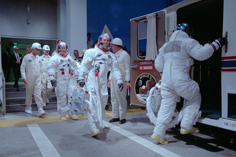 Three astronauts wearing spacesuits and group of engineers exit building and walking towards van.