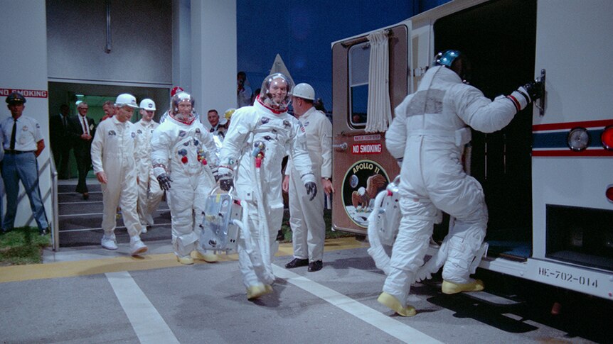 Three astronauts wearing spacesuits and group of engineers exit building and walking towards van.