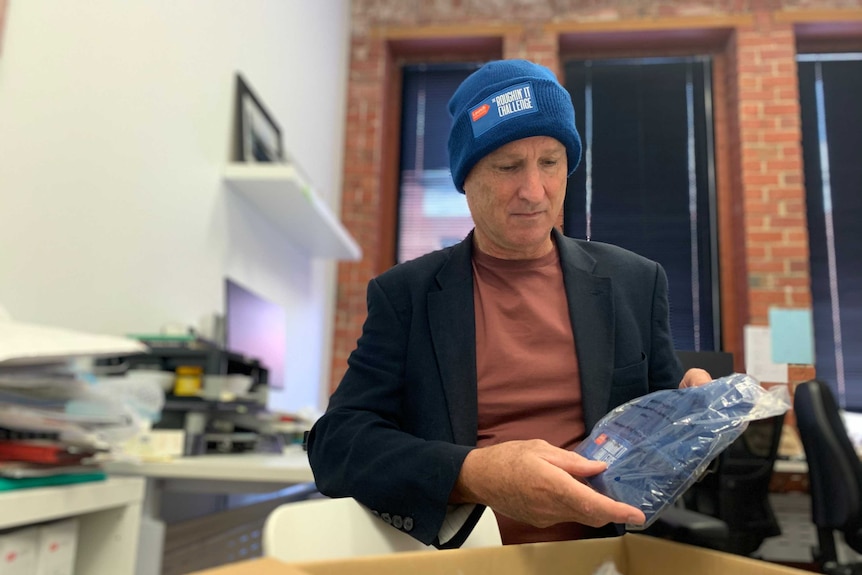 A man wearing a blue beanie holds another beanie in his hands, above a cardboard box.