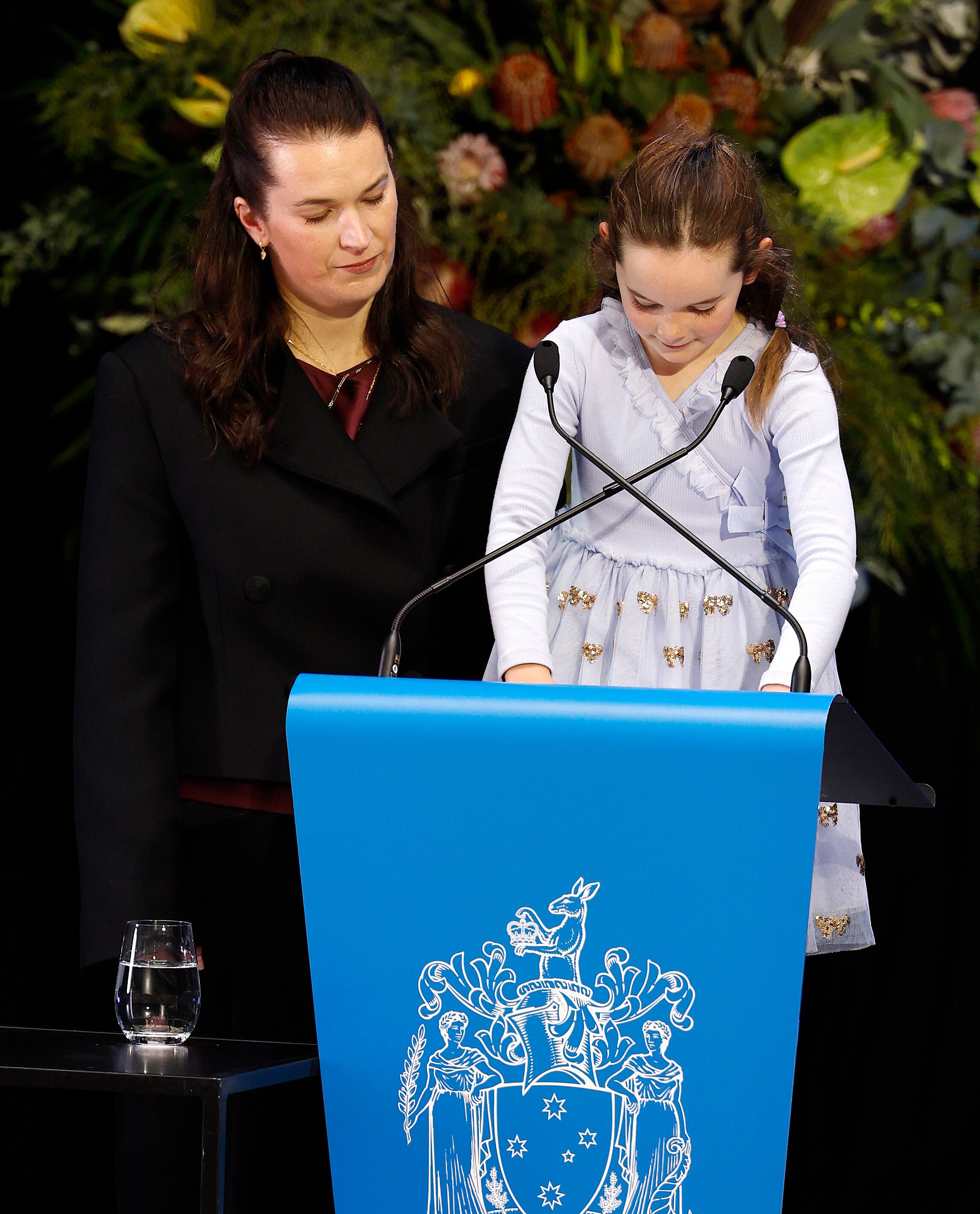 A woman with long dark hair stands at a lectern beside a young girl who reads into a microphone.