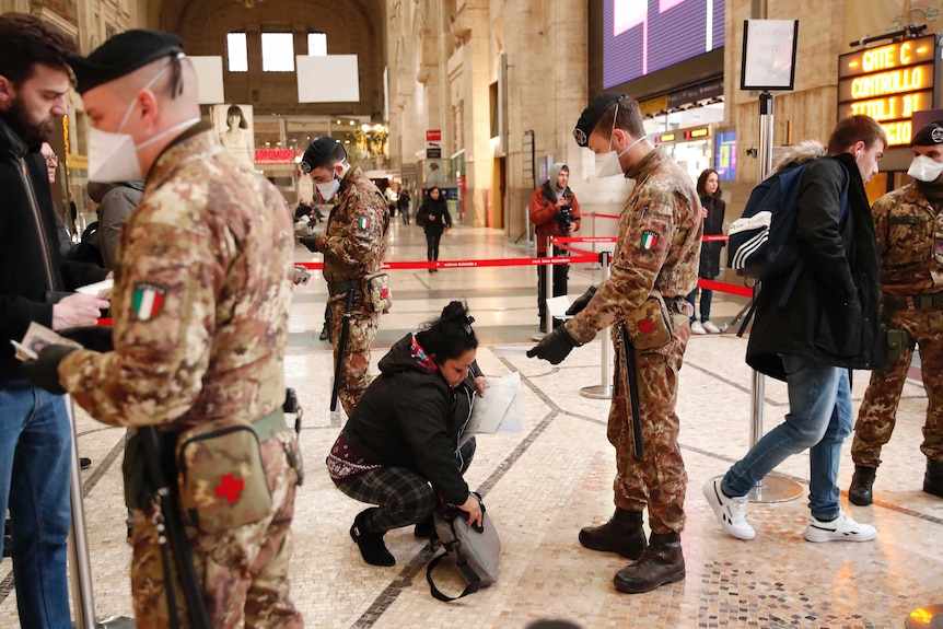 A woman bends down to remove something from her bag while a soldier in a face mask watches on.