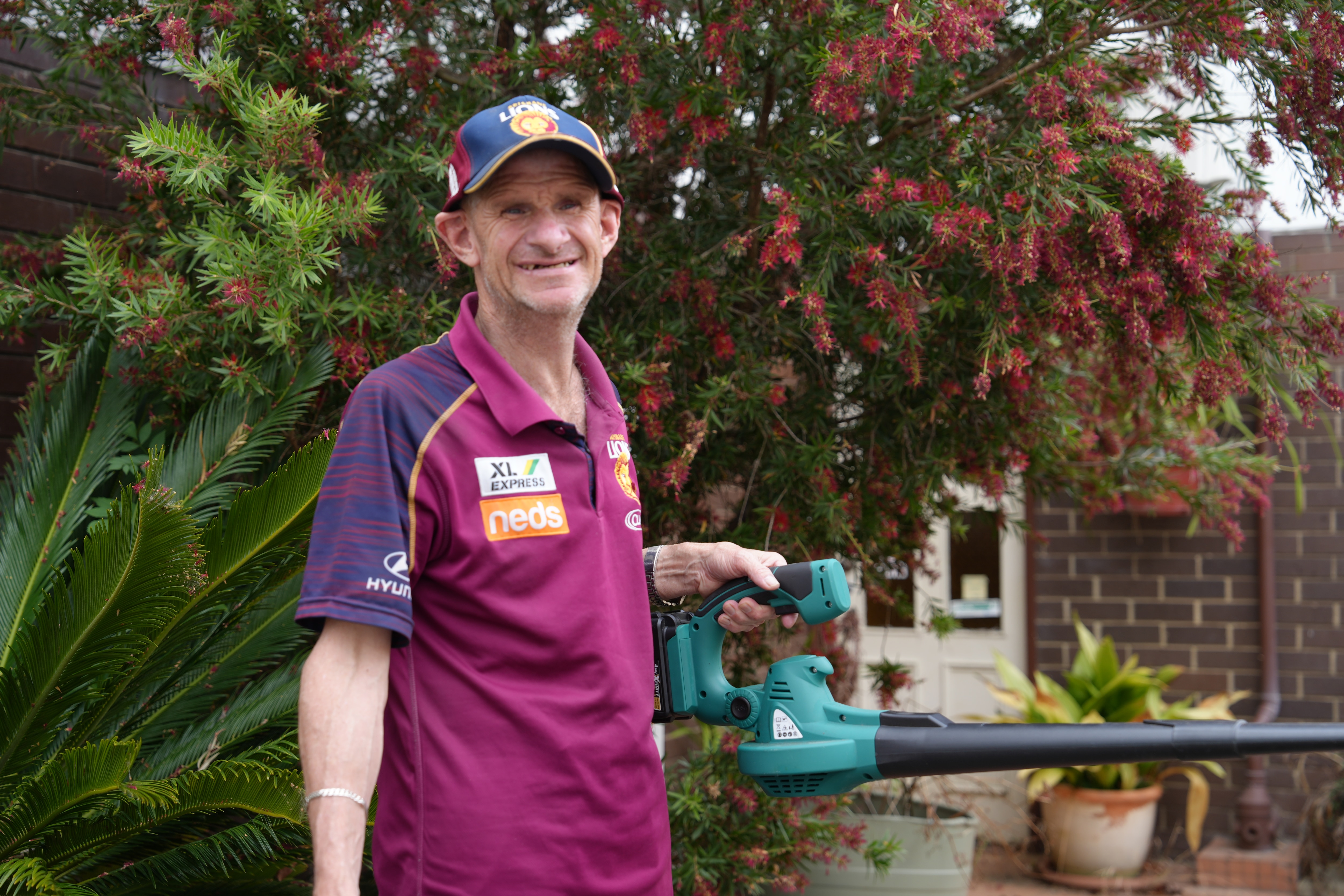 A man in a Brisbane Lions branded jersey and cap smiles with a leaf blower in his left hand.