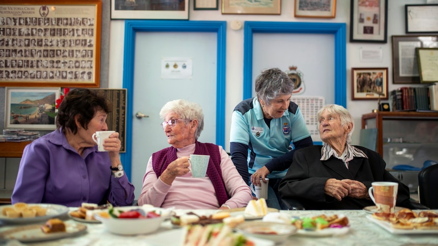 Four older women chat over cups of tea with sandwiches on a table in front of them and ANZAC posters framed behind them.