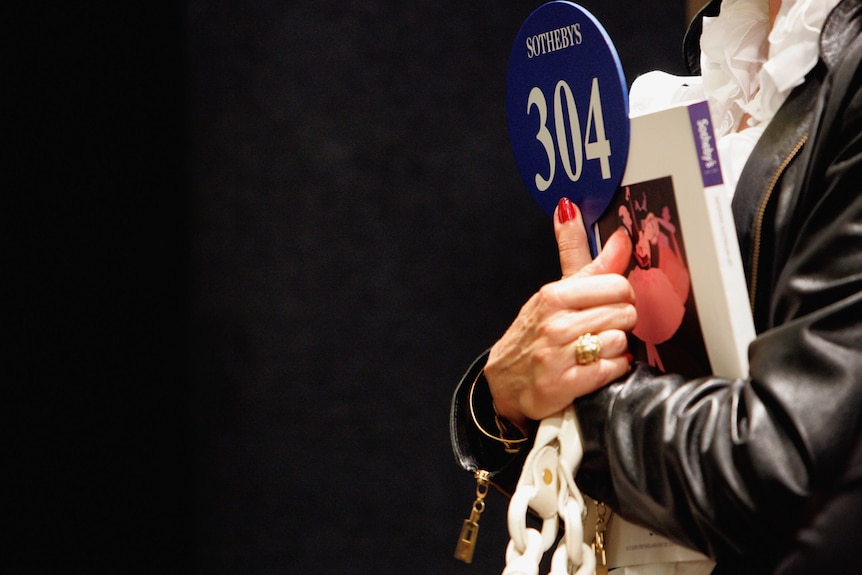 A person holds an art catalogue and bidding paddle close to their chest with their arms crossed.