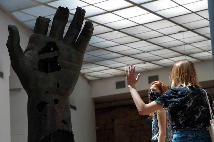 You view two women in masks staring up at a large ancient bronze statue of a hand in a bright museum room.