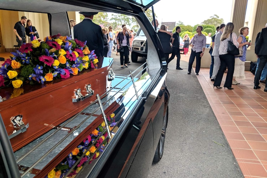 A coffin in the back of a long car, with flowers on it.