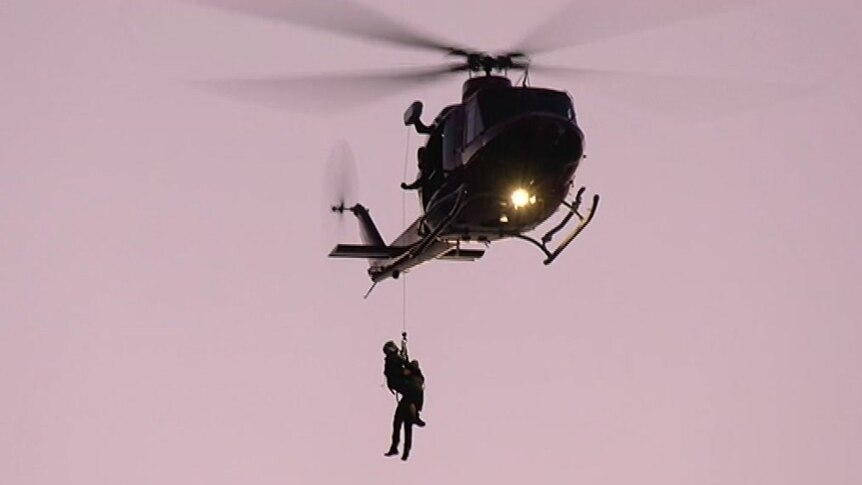A helicopter with two people hanging from a rope with a pink sky background