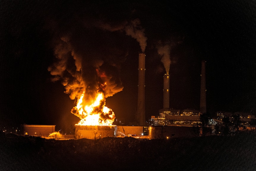 Flames rise from an oil tank on fire next to two smoking chimneys at night.