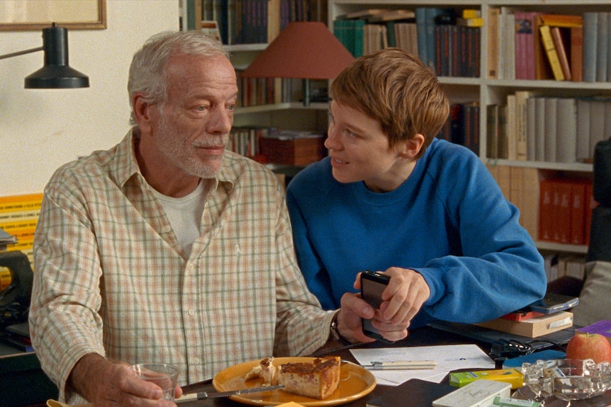 An older man sits at a table eating a slice of cake, looking blank as a younger woman in a blue jumper talks to him.