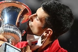 Novak Djokovic kisses a silver trophy with a white face mask around his chin
