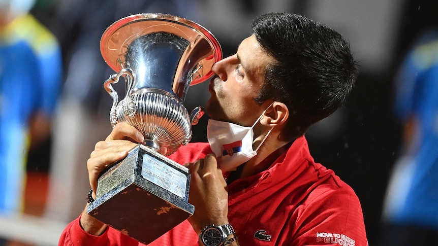 Novak Djokovic kisses a silver trophy with a white face mask around his chin