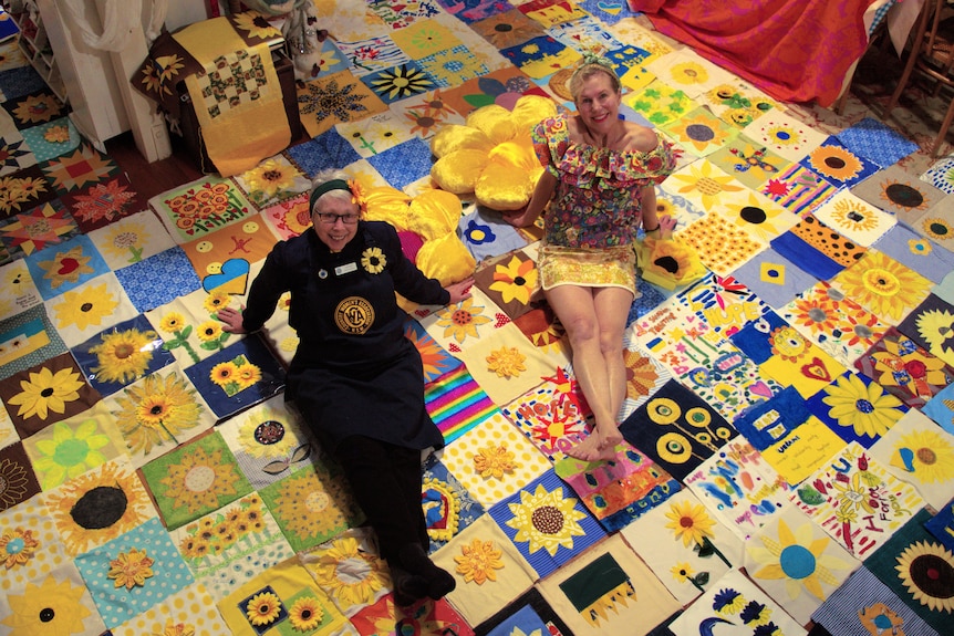 two women sit on a sunflower themed quilt and look to the camera above them