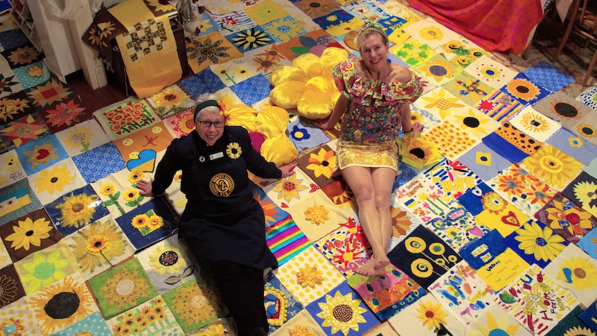 two women sit on a sunflower themed quilt and look to the camera above them