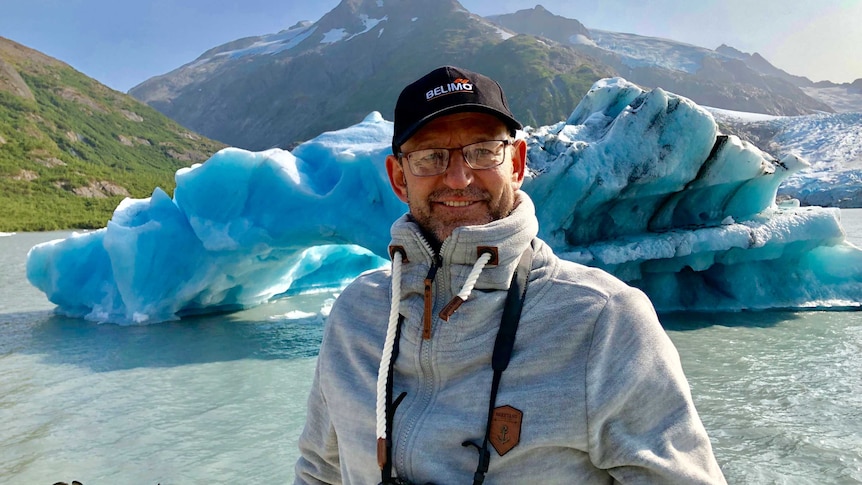 A man smiles in foreground, iceberg and hills in the background.