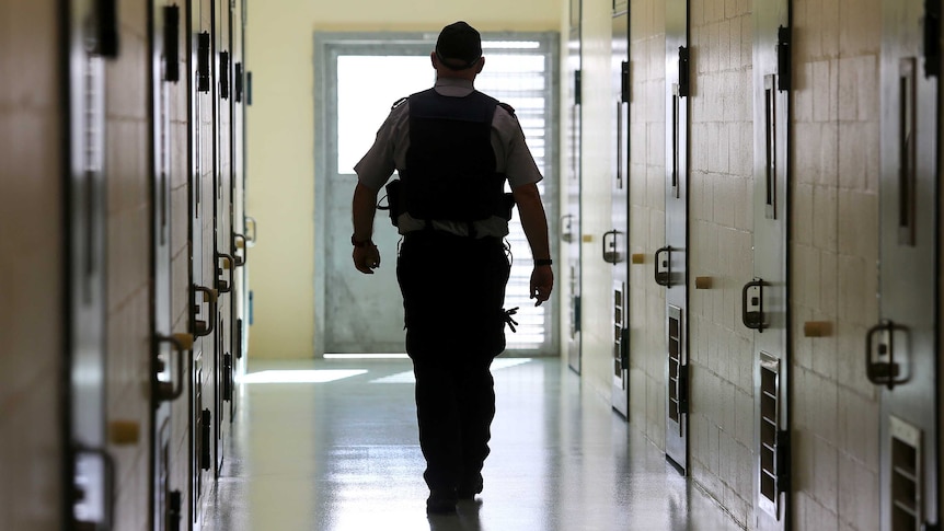 A silhouetted corrections officer walks down a cell corridor in a prison.