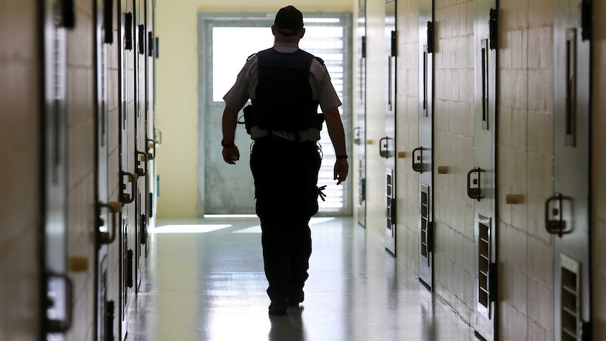 A silhouetted corrections officer walks down a cell corridor in a prison.