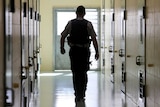 A silhouetted corrections officer walks down a cell corridor in a prison.