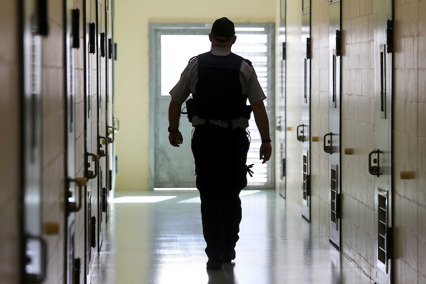 A silhouetted corrections officer walks down a cell corridor in a prison.