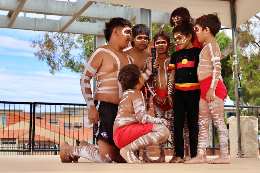 Six young Indigenous boys wearing white body paint gather round a microphone on a stage.