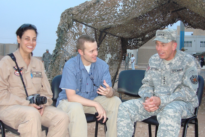 A man in a US Army uniform sits on a chair with a journalist seated next to him chatting. A woman in Army attire sits left