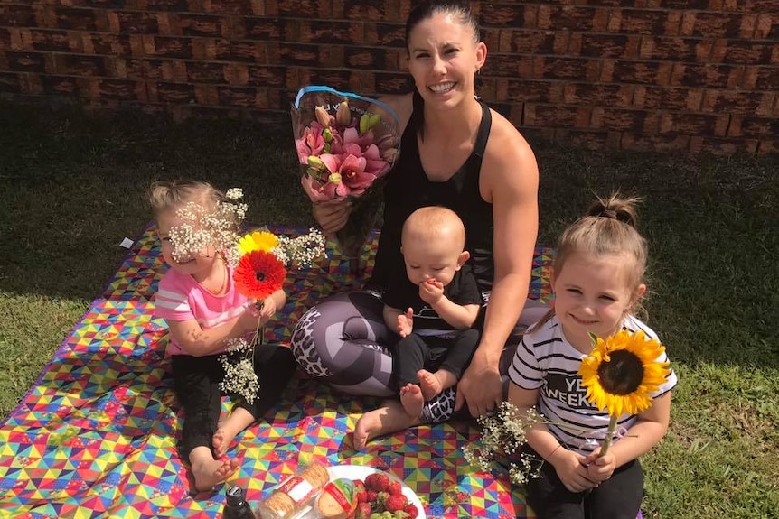 Hannah Clarke sits with her three children on a picnic rug with a fruit platter and holding flowers.