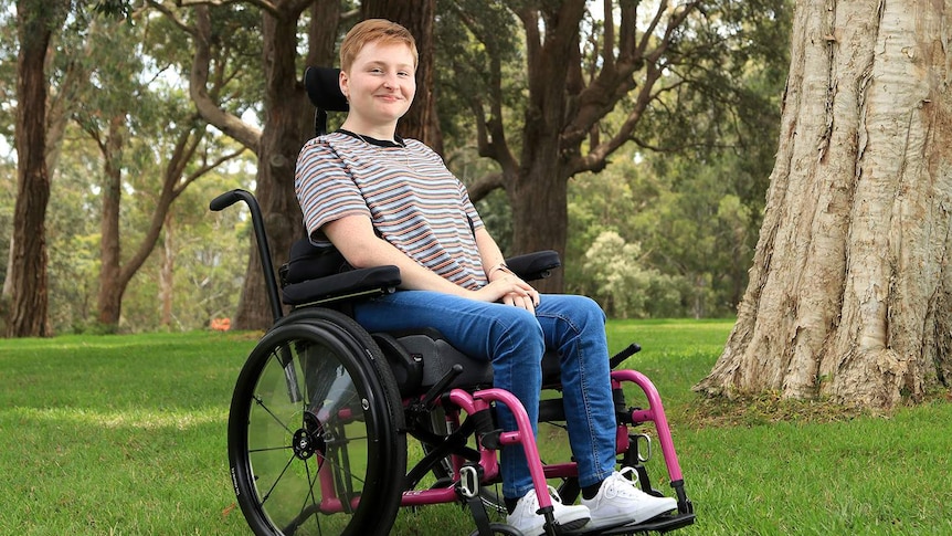 A photo of Lily sitting on a wheelchair in a park, she has a smile on her face