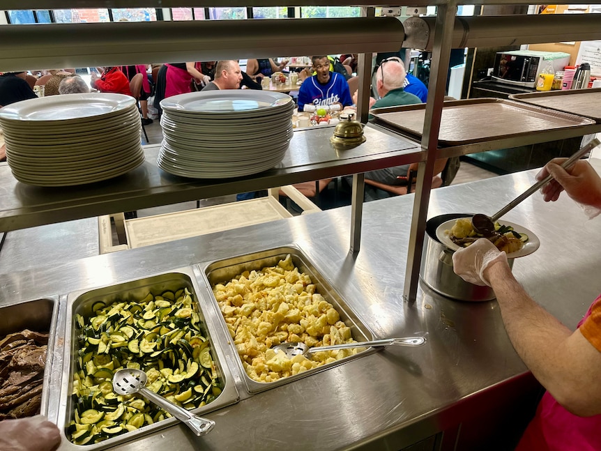 A cafeteria bench, stacks of plates and tubs of food being served up. 