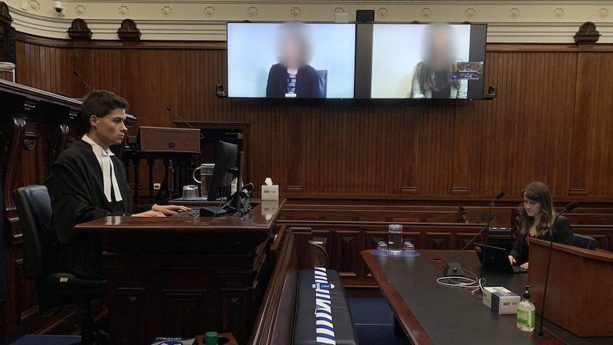 A wooden court room is in picture. Two staff sit at a distance from each other, with two people on screens.