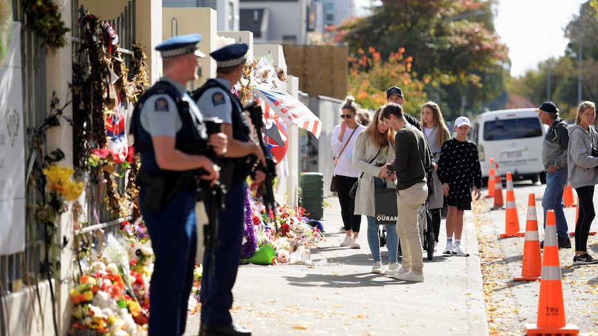 Police stand guard at the entrance of Al Noor Mosque, which is still covered in flowers and cards.