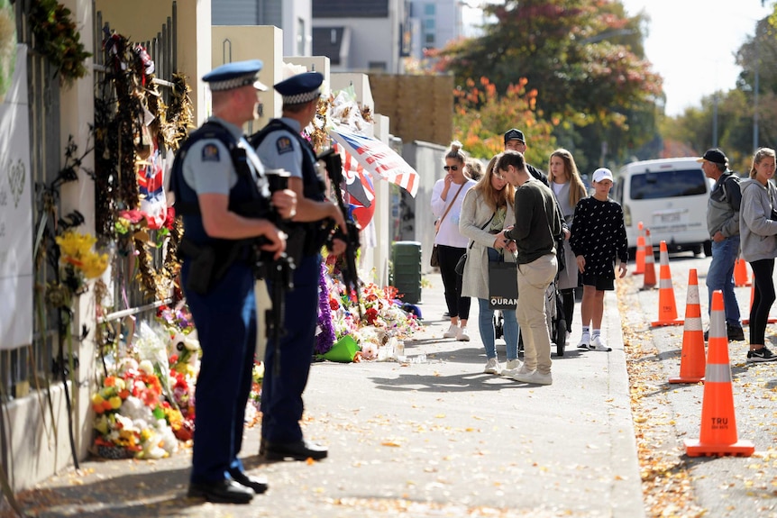 Police stand guard at the entrance of Al Noor Mosque, which is still covered in flowers and cards.