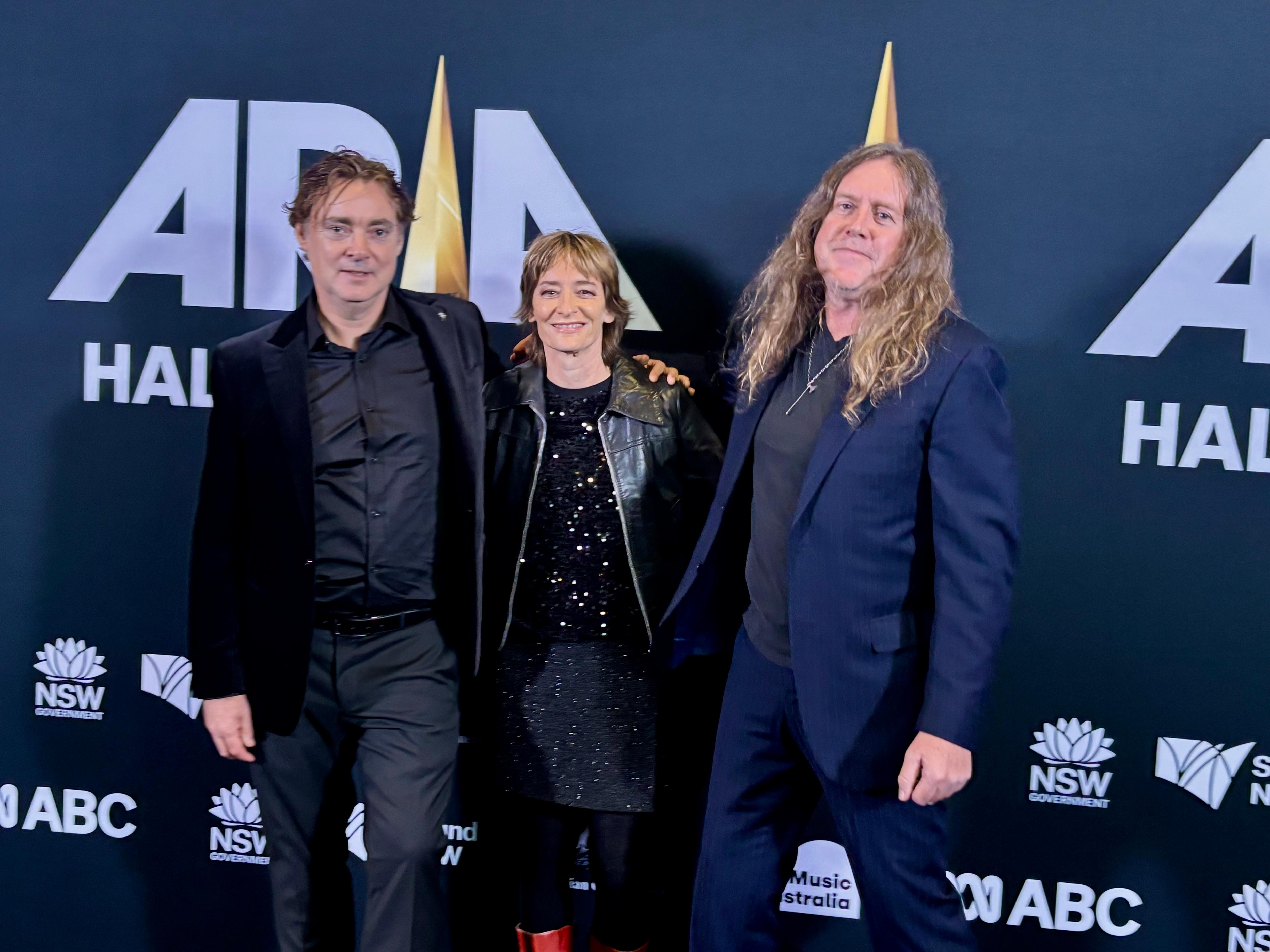 Two long-haired men flank a shorter and younger woman at an awards' night