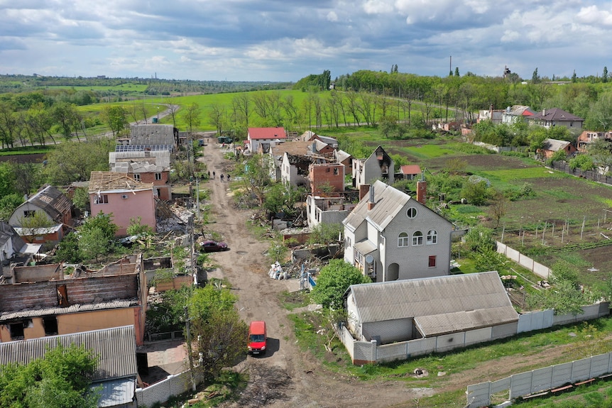 An aerial shot shows partially destroyed houses in a small village. Some roofs have been ripped off or caved in