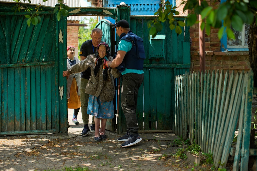 an elderly woman in a head scarf is led through a blue fence by two men in blue 