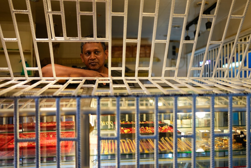 A man looking through a caged window with display shelves underneath him. 