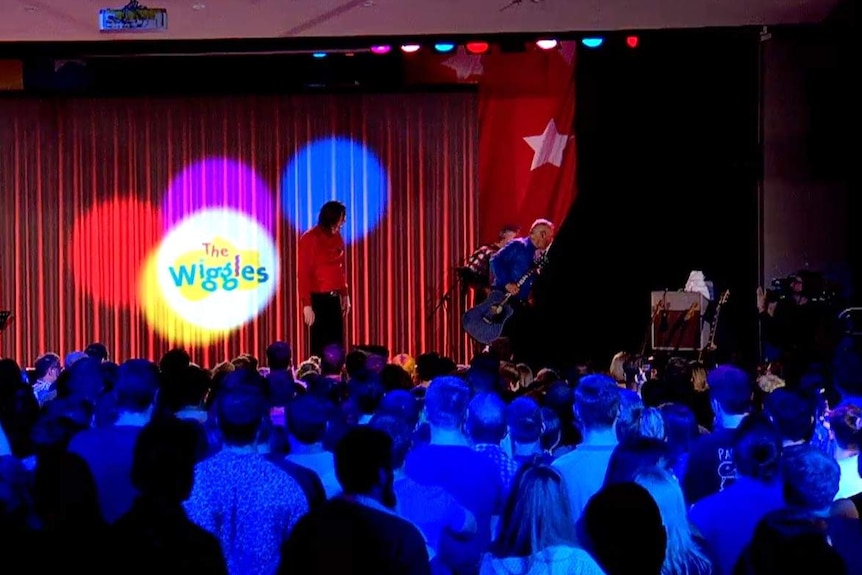 a stage with a red curtain and wiggles logo with the red and blue wiggles looking to their left where a black curtain is drawn