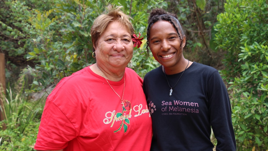 Two women smile at the camera with greenery behind