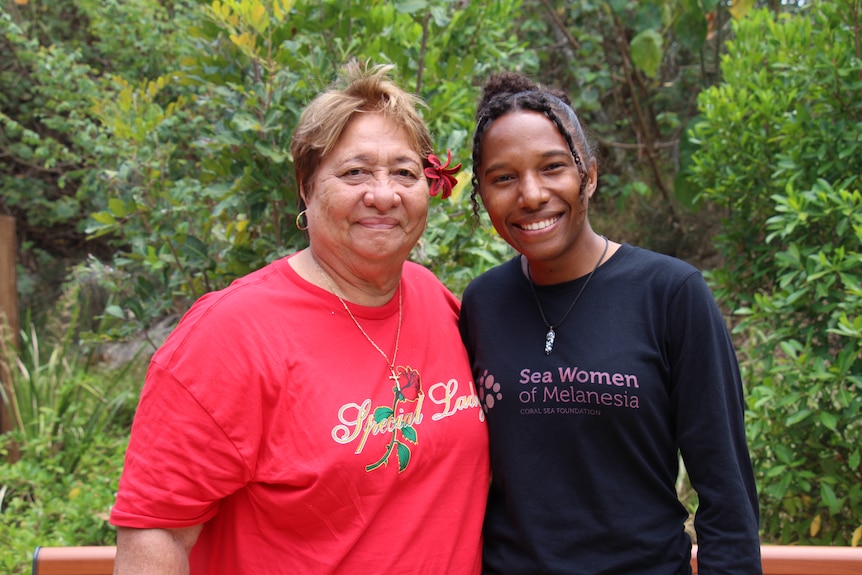 Two women smile at the camera with greenery behind