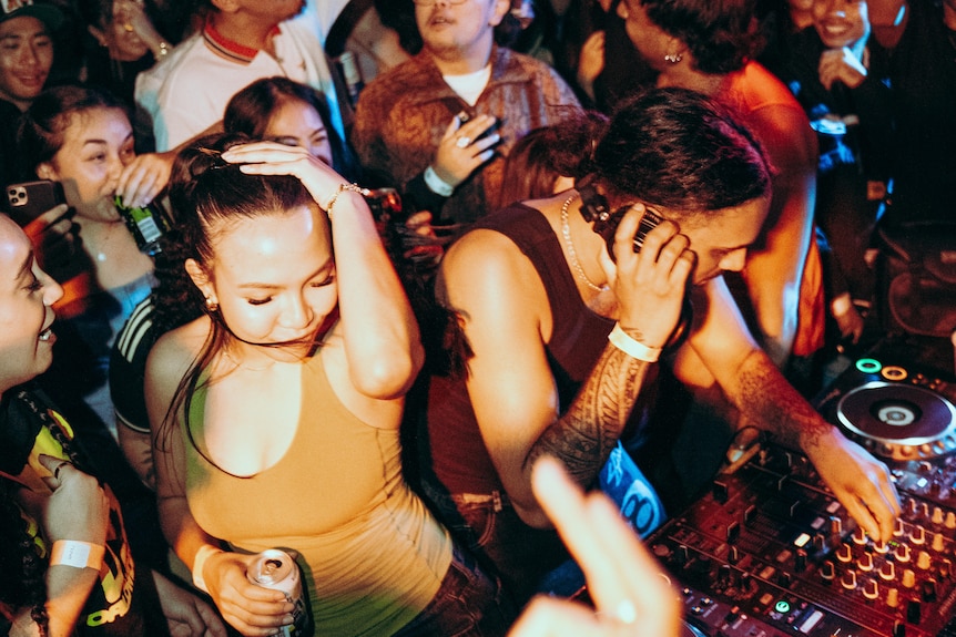 A crowd of young people mosh behind a DJ. 