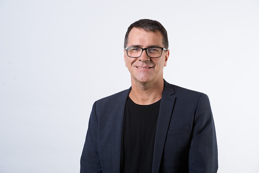 A man in a black shirt and dark blue blazer wearing glasses stands in front of a white background.