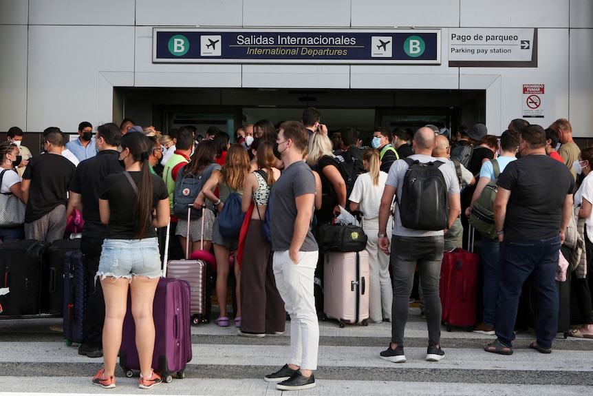 A crowd of people with luggage and backpacks stand near the entrance to an airport building.
