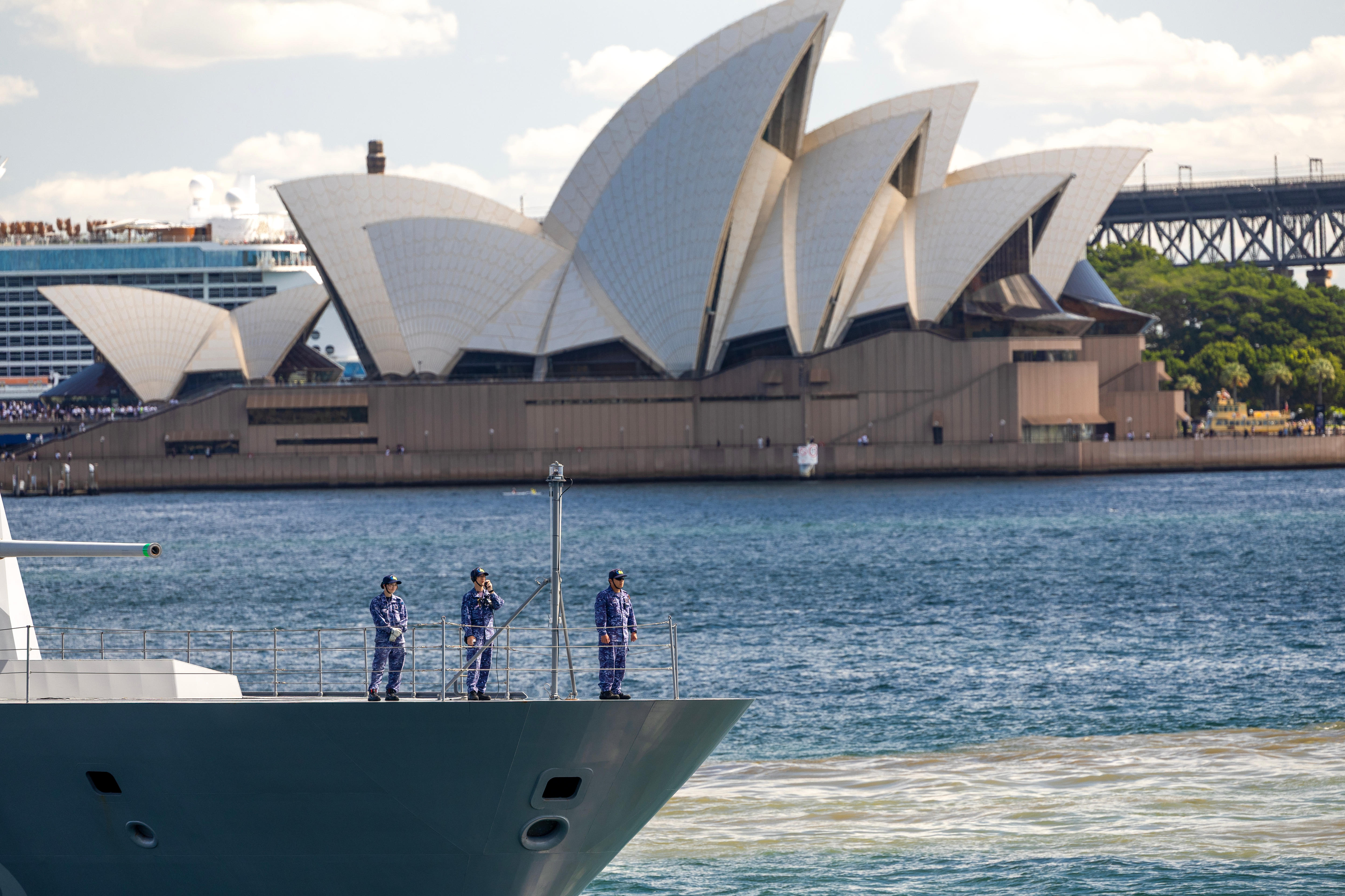 Tres hombres uniformados se encuentran en la cubierta de una fragata gris mientras zarpa, con la Ópera de Sydney detrás.