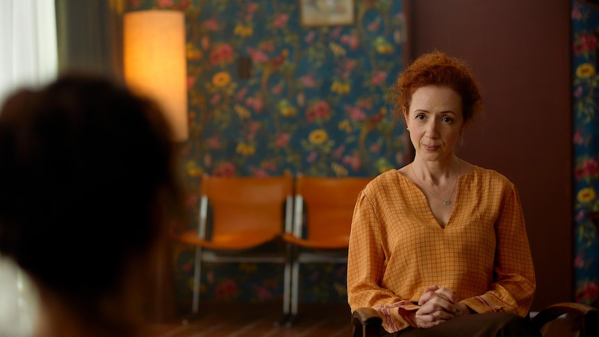 White woman with fiery red curly hair wears a mustard blouse and sits opposite a patient in a therapist's office.